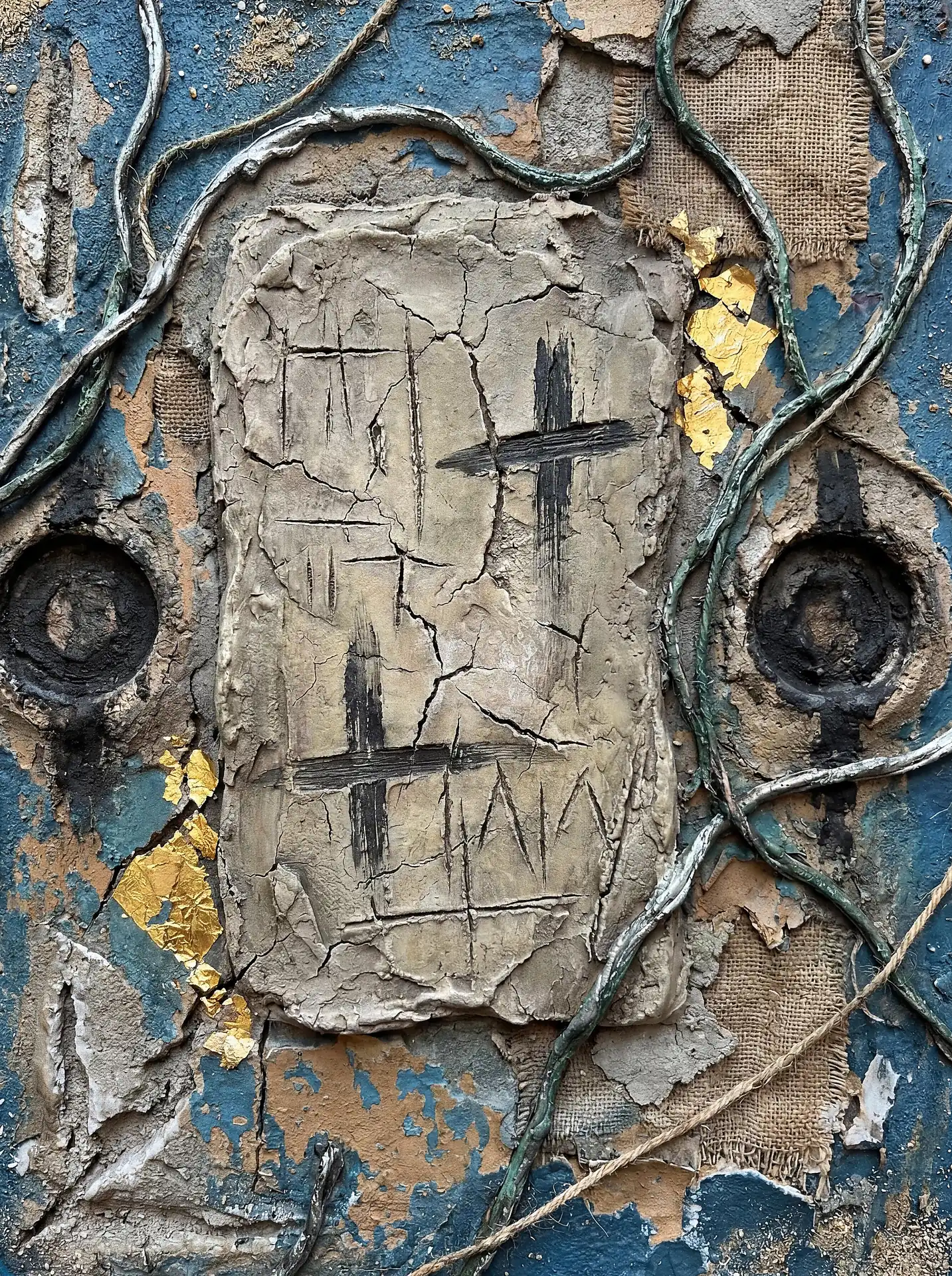 Deep blue tones and golden highlights illuminate this textured abstract relief featuring a central cracked clay slab with dark primitive markings. The composition incorporates metallic vines, burlap patches, and peeling paint to create a rustic, archaeological aesthetic. Artwork by Constantine Cheshire