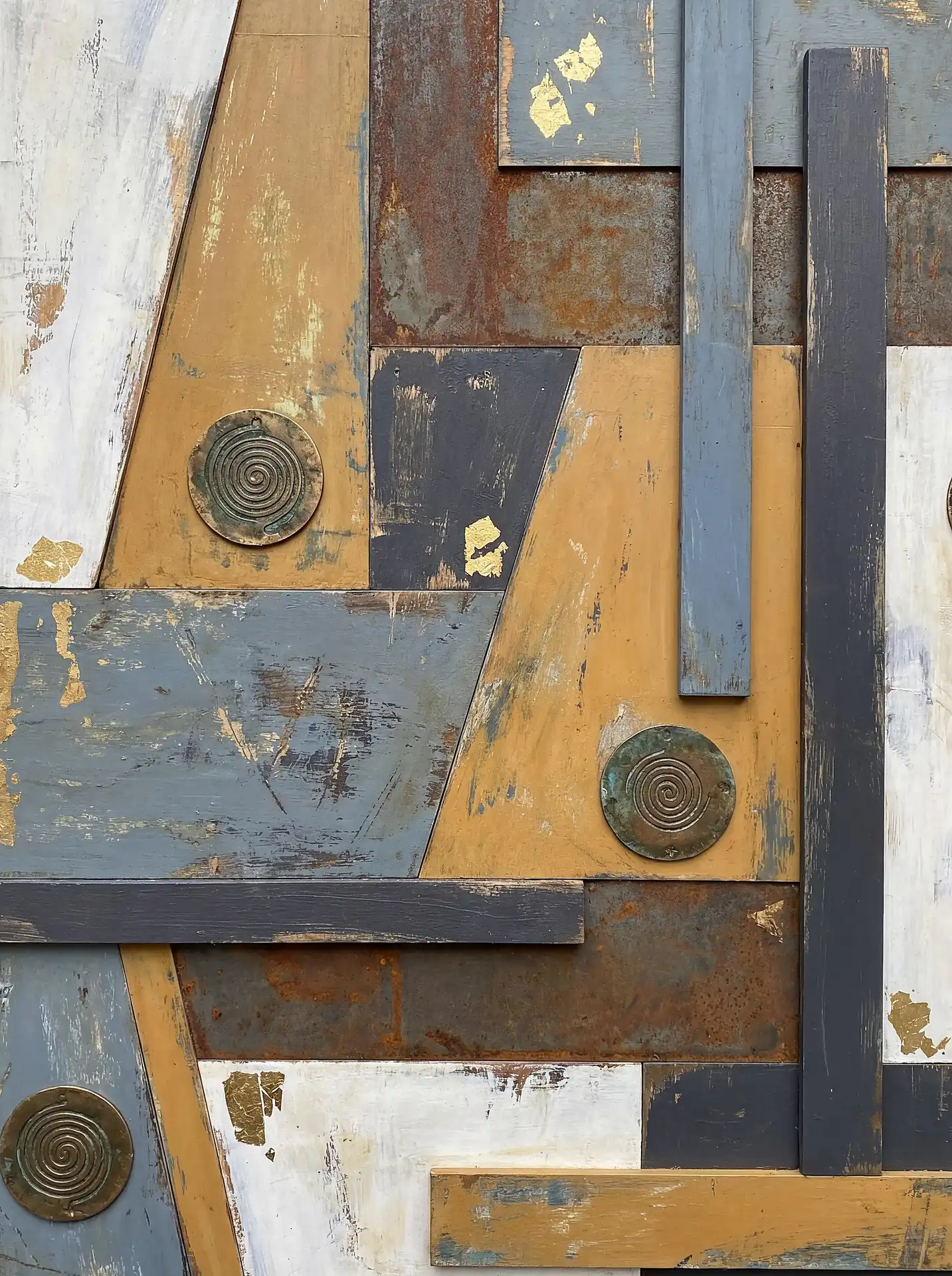 Weathered wood, rusted metal, and shimmering gold leaf form a complex geometric assemblage featuring circular bronze spirals. This contemporary industrial artwork combines cool blue and white tones with warm ochre and metallic accents for a rugged, textured feel. Artwork by Constantine Cheshire