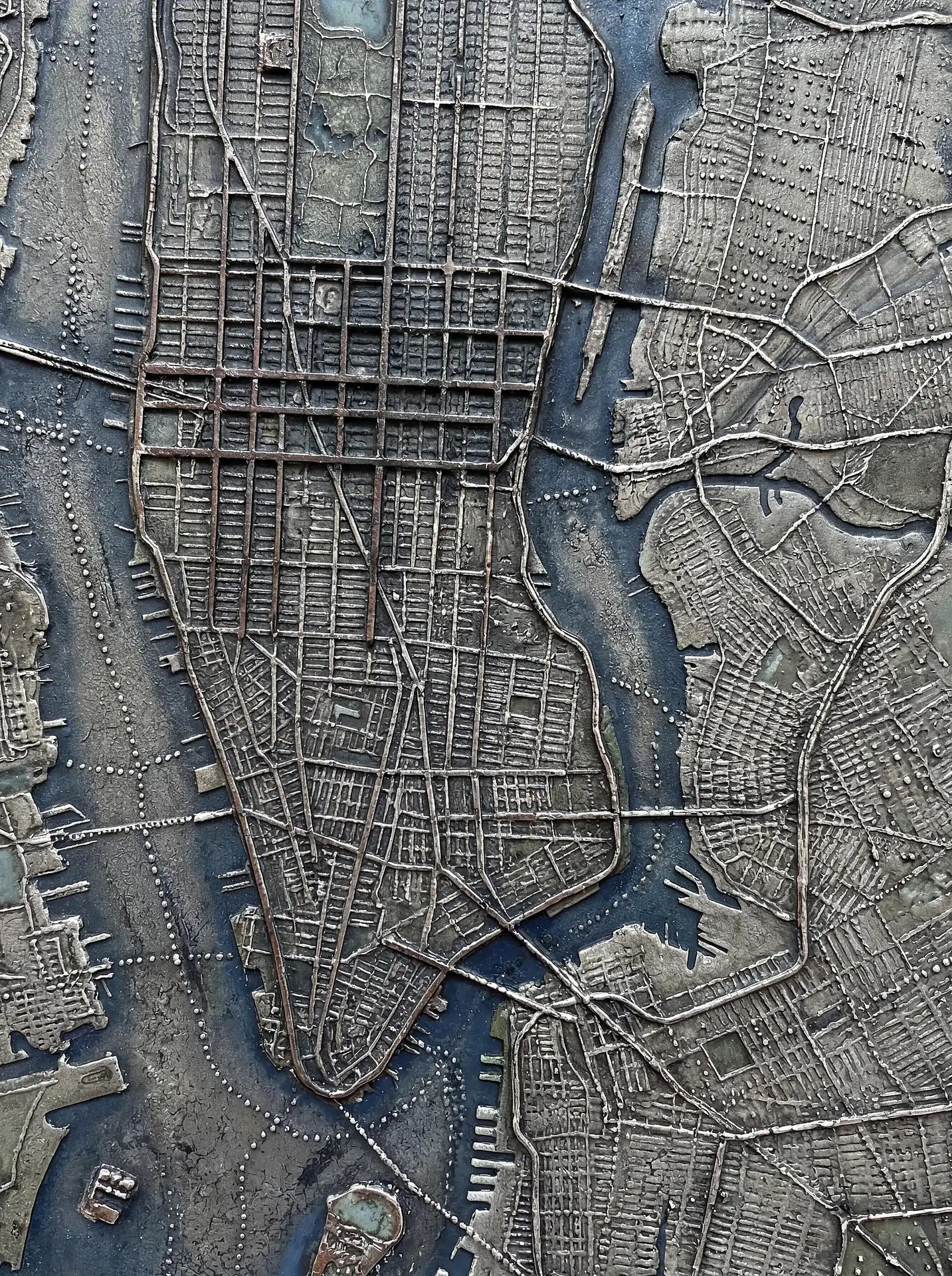 Muted silver and bronze tones define this relief map of Manhattan, showing the dense grid of New York City streets. This industrial metallic sculpture features raised textures for landmasses and a deep blue hue for the surrounding rivers. Artwork by Constantine Cheshire