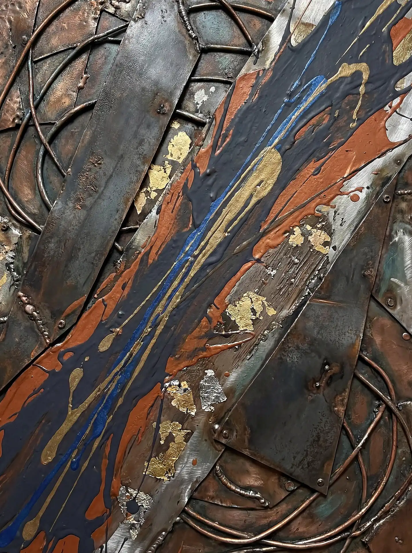 Layering of weathered metal plates and coiled copper wire creates a complex industrial foundation for vibrant splashes of blue and orange paint. This abstract mixed-media piece features gold leaf accents and welded textures, offering a rugged yet sophisticated contemporary aesthetic in earthy tones. Artwork by Constantine Cheshire