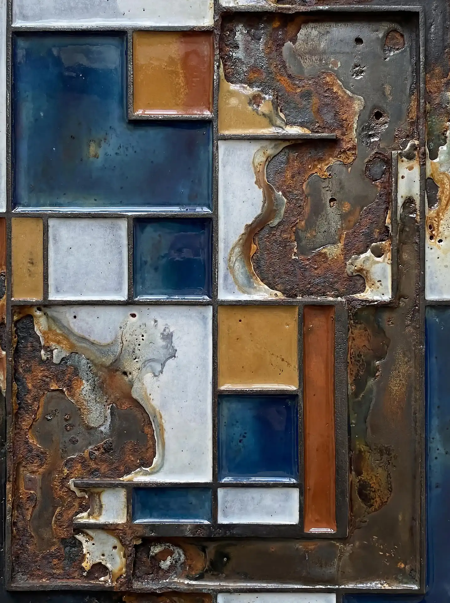 Weathered metal and glossy enamel tiles form a geometric grid of contrasting textures and colors. This industrial abstract composition pairs deep blues and oranges with heavy oxidation and pitted surfaces to explore the intersection of order and decay. Artwork by Constantine Cheshire