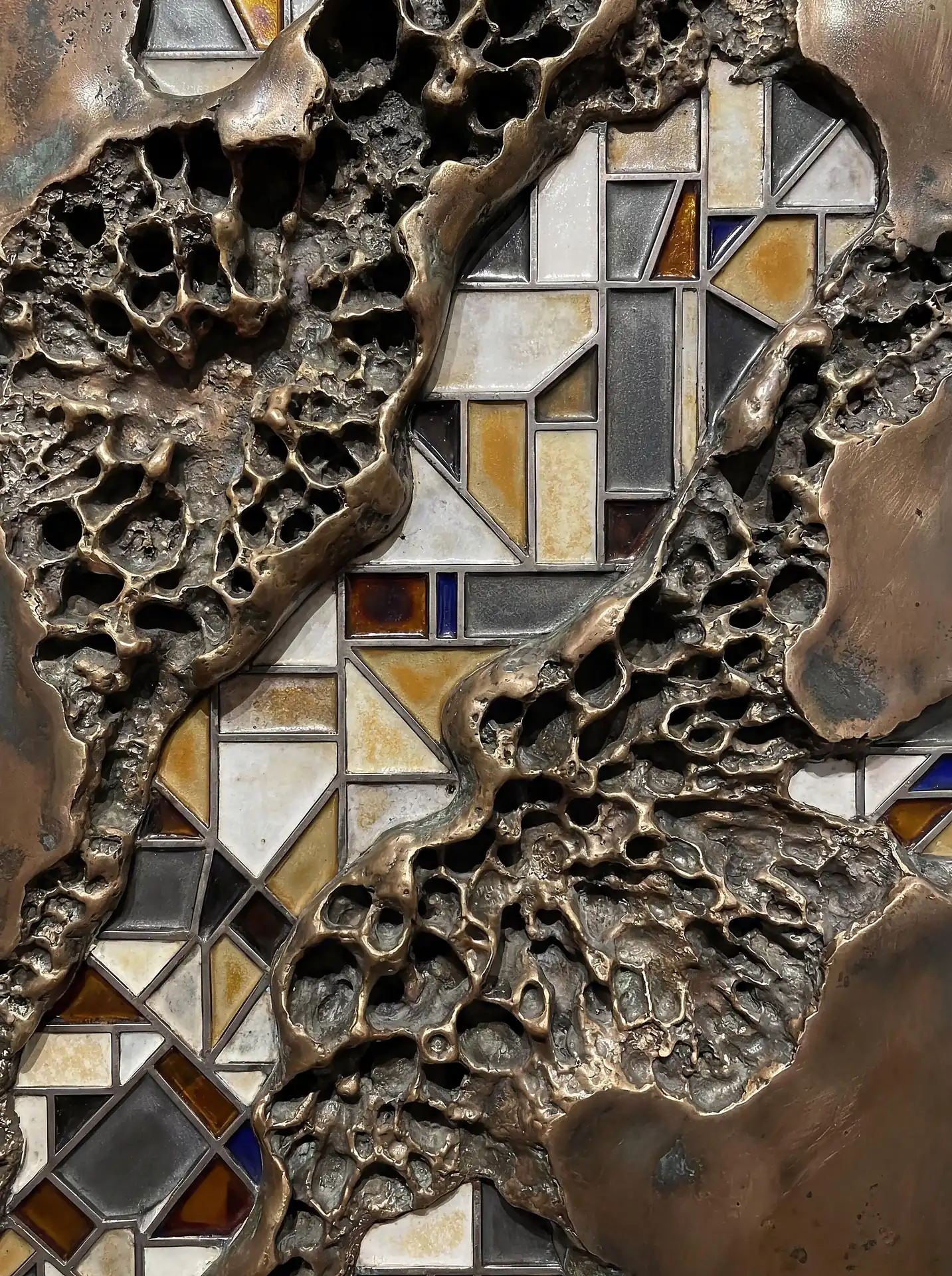 Flowing across the frame, textured molten metal fragments reveal a precise geometric glass mosaic underneath. This mixed-media artwork combines organic, porous bronze surfaces with sharp-edged tiles in muted tones of amber, white, and grey for a sophisticated industrial aesthetic. Artwork by Constantine Cheshire