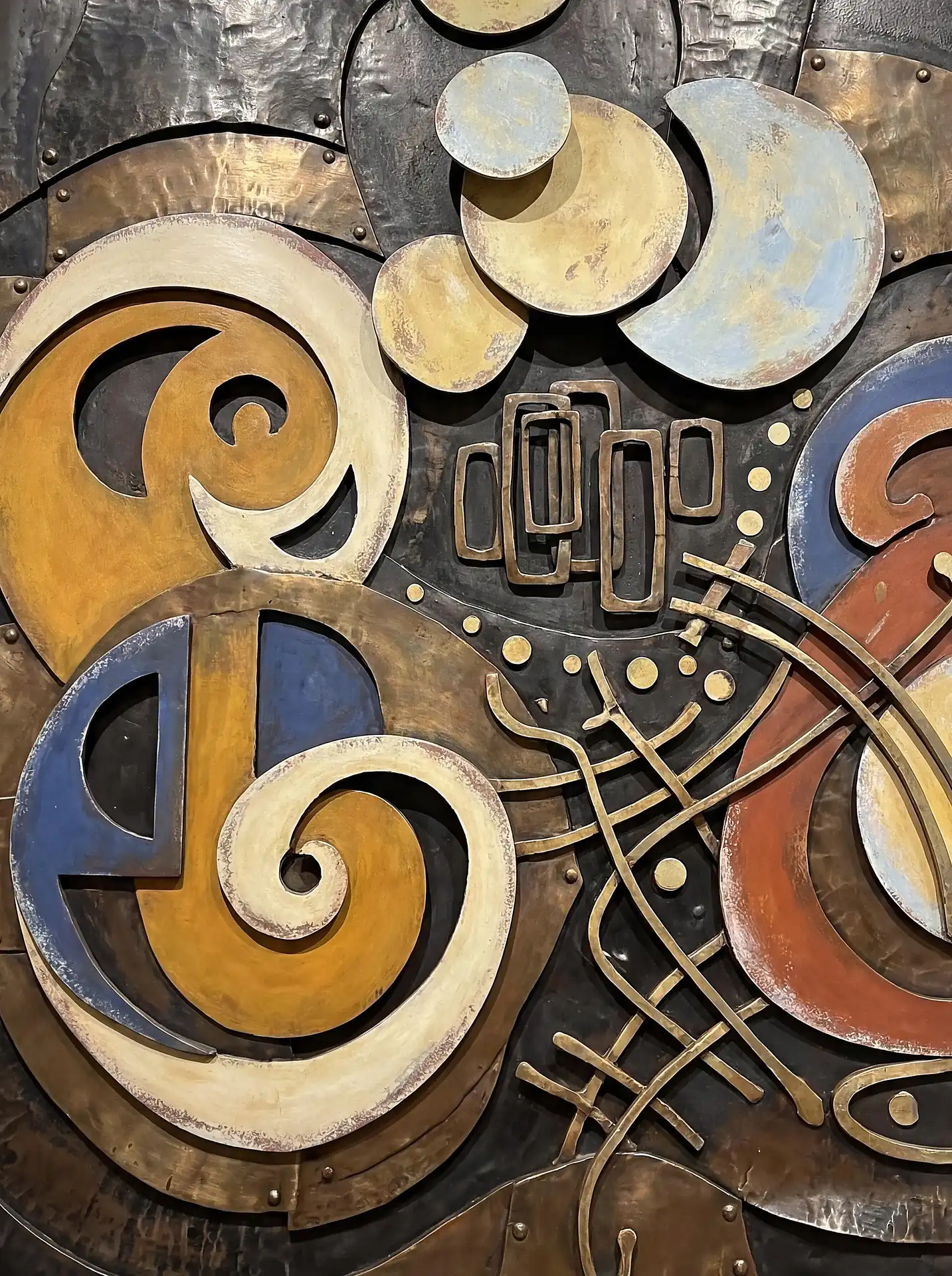 Sophisticated and industrial, this mixed-media relief sculpture features weathered copper spirals and hammered brass plates secured by delicate rivets. The composition balances geometric rectangles with organic circular forms in a rich, aged palette of bronze, blue, and gold. Artwork by Constantine Cheshire