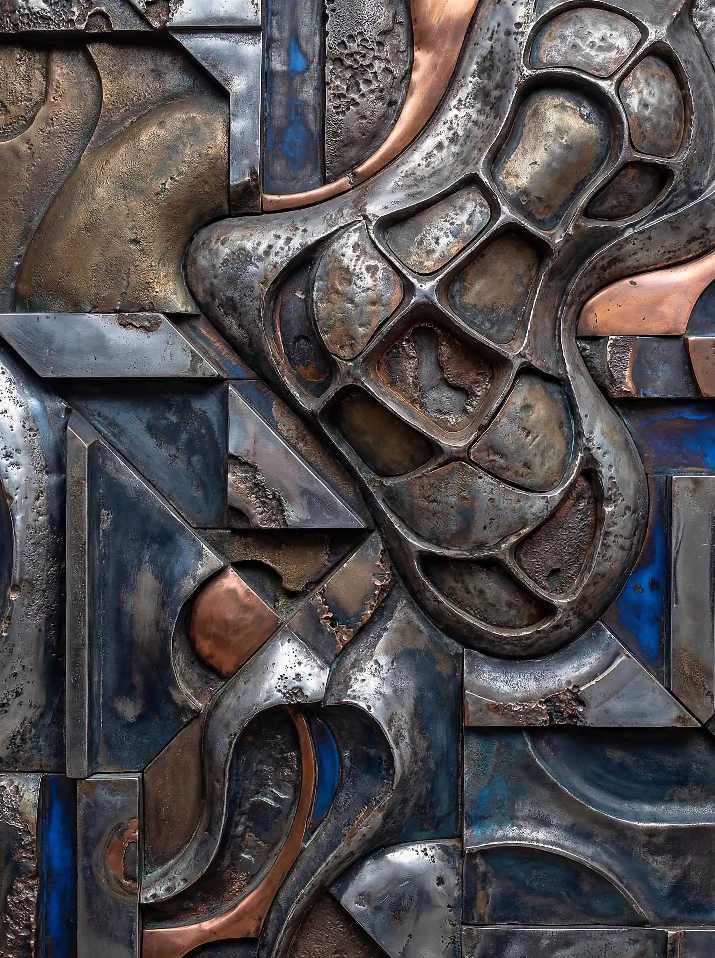 Cool blue and warm copper tones highlight this abstract metal relief sculpture made of weathered steel and iron plates. The industrial artwork combines heavy metallic textures with organic and geometric forms to create a complex, modern wall piece. Artwork by Constantine Cheshire